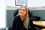 Jane Swift, a light-skinned woman with long gray hair wearing a black shirt, sits at her desk at the Education at Work headquarters in Arizona.